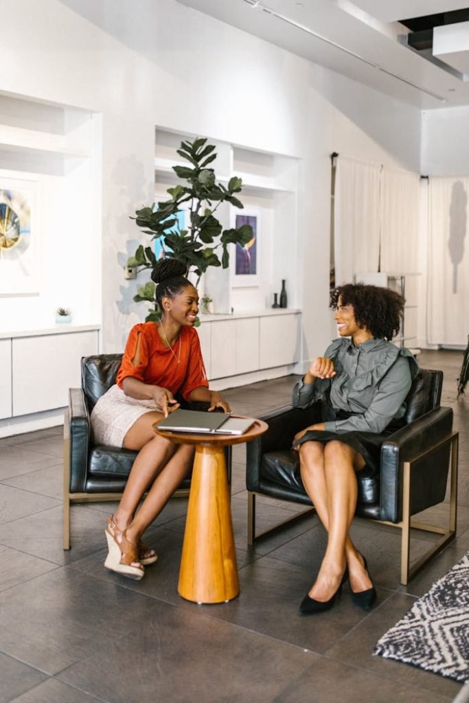 Two women in an office setting sharing a friendly conversation, smiling and interacting warmly.